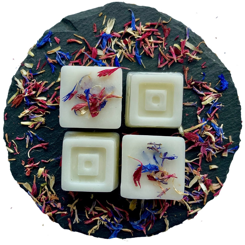 Four square-shaped Maldives-scented wax melts arranged on a circular grey slate decorated with multi-coloured cornflowers.