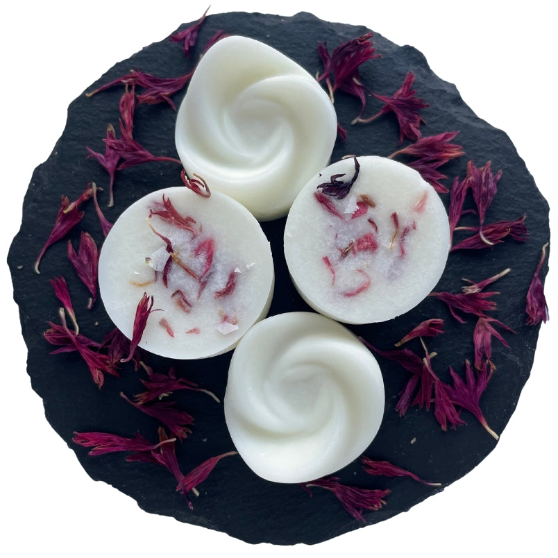 Four swirl-shaped peach and pear scented wax melts on a circular slate with pink cornflower botanicals as decoration.