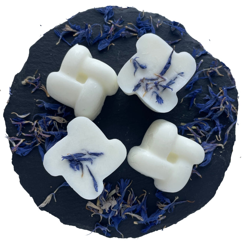 Four knot-shaped rock salt and driftwood scented wax melts arranged on a circular slate with blue cornflower botanicals.